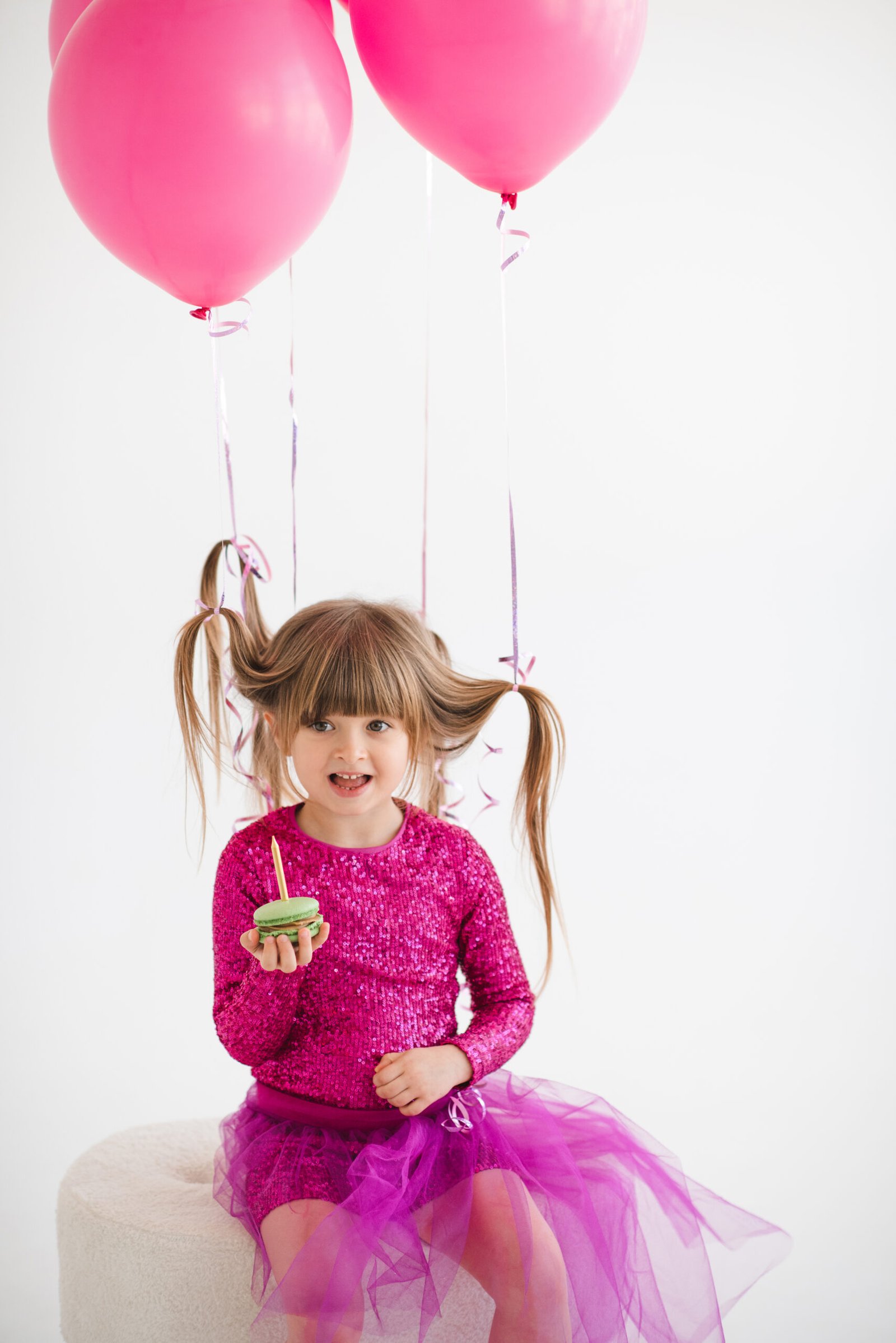 Cute happy child girl 4-5 year old wear princess dress posing with balloons eating cake with candle having birthday party over white in room. Childhood.
